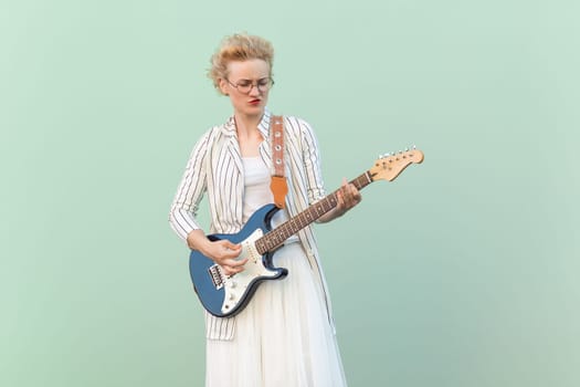 Portrait of blonde attractive young adult hipster woman in striped shirt and white skirt with eyeglasses plating guitar like star. Indoor studio shot isolated on light green background.