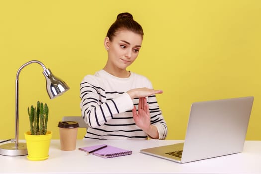 Tired woman manager showing time out gesture looking at laptop screen, talking on video call, asking pause, break of online communication. Indoor studio studio shot isolated on yellow background.