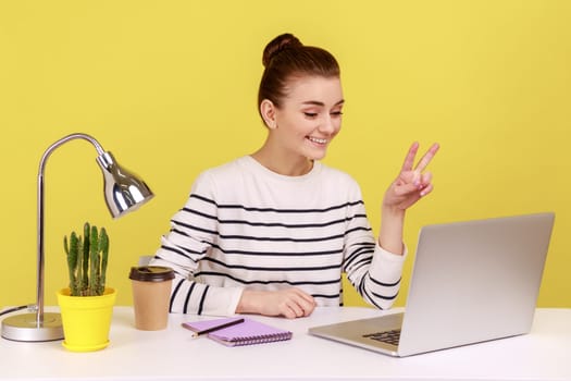 Positive optimistic woman office manager showing victory gesture and smiling looking into laptop screen, talking video call, online conference. Indoor studio studio shot isolated on yellow background.
