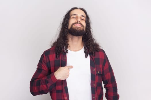 This is me. Satisfied proud bearded man in checkered red shirt points at himself, stands self confident, feels successful of his own achievement. Indoor studio shot isolated on gray background.