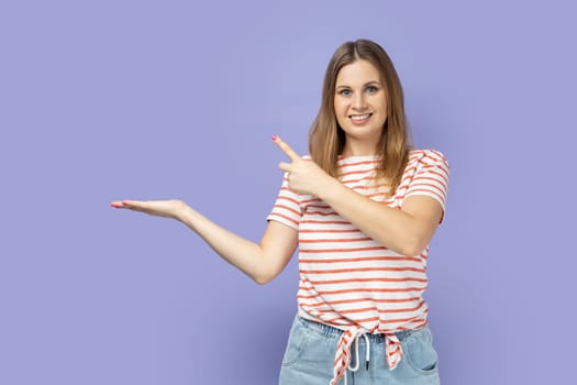 Portrait of blond woman wearing striped T-shirt presenting advertising area on her palm and pointing to copy space, holding empty place for commercial. Indoor studio shot isolated on purple background