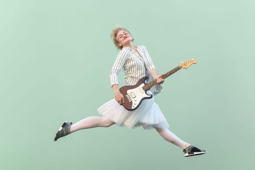 Portrait of young adult happy blonde woman in white shirt, skirt, and striped blouse with eyeglasses holding electric guitar, smiling and jumping. Indoor studio shot isolated on light green background