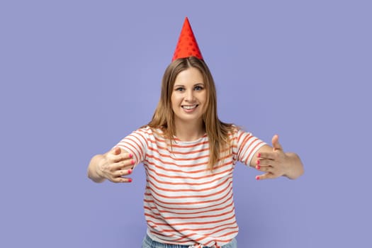 Portrait of adorable hospitable woman in striped T-shirt and party cone, celebrating her birthday, smiling and reaching out hands, going to embrace. Indoor studio shot isolated on purple background.