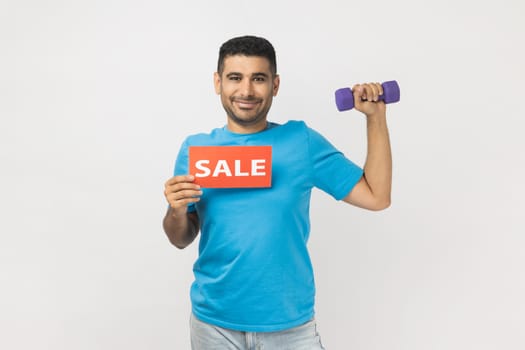 Portrait of smiling delighted man wearing blue T- shirt standing holding dumbbell and card with sale inscription, discounts for fitness season ticket. Indoor studio shot isolated on gray background