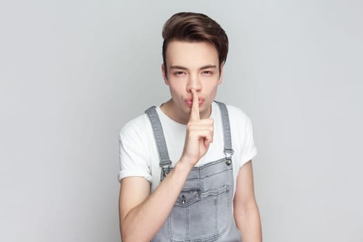 Portrait of mysterious brunette man standing presses index finger to lips, makes hush gesture tells secret, asks to be quiet, wearing denim overalls. Indoor studio shot isolated on gray background.