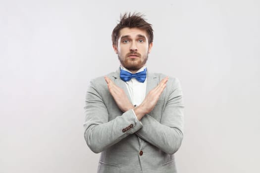 Portrait of upset attractive bearded man standing showing x sign, no way gesture, looking at camera, wearing grey suit and blue bow tie. Indoor studio shot isolated on gray background.