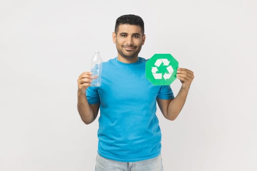 Portrait of smiling happy handsome unshaven man wearing blue T- shirt standing holding green recycling sign, planet protection. Indoor studio shot isolated on gray background.