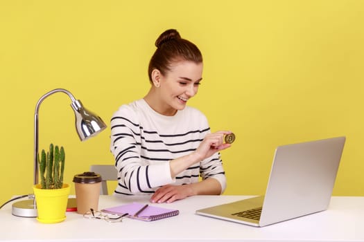 Cryptocurrency, digital money. Positive woman sitting in office workplace, showing golden bitcoin to laptop screen, smiling to display. Indoor studio studio shot isolated on yellow background.