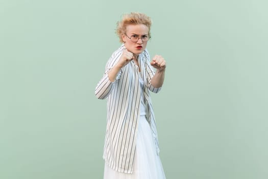 Portrait of angry aggressive attractive blonde woman wearing striped shirt and skirt, clenching fists, being ready to attack. Indoor studio shot isolated on light green background.