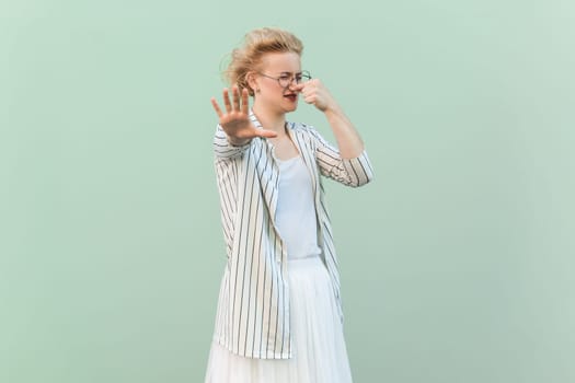 Portrait of confused disgusted blonde woman wearing striped shirt and skirt pinching her nose, showing stop gesture. Indoor studio shot isolated on light green background.