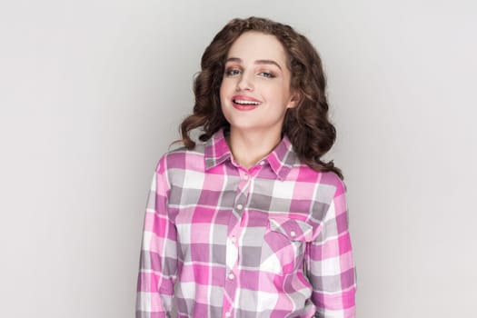Portrait of woman with curly hair being very glad smiling with broad smile showing her perfect teeth, having fun, wearing pink checkered shirt. Indoor studio shot isolated on gray background.