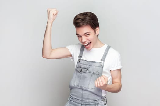 Yes, finally success. Brunette man raises arms with clenched fists, celebrates something has upbeat mood, feels euphoric, wearing denim overalls. Indoor studio shot isolated on gray background.
