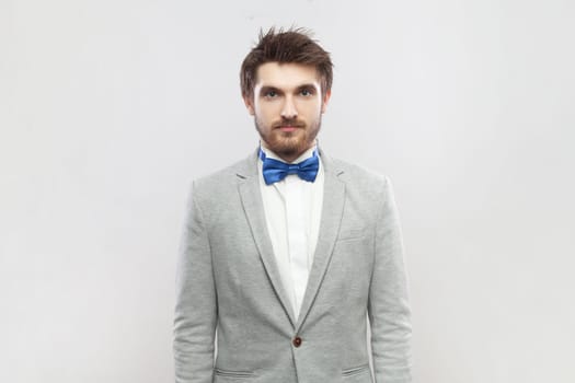 Portrait of calm positive bearded man standing looking at camera, having relaxed facial expression, wearing grey suit and blue bow tie. Indoor studio shot isolated on gray background.