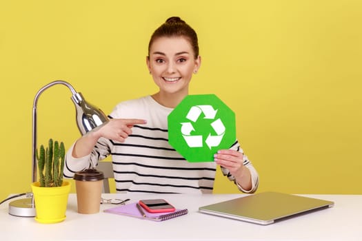 Positive satisfied woman pointing at green recycling symbol in her hands, ecology concept, sitting on workplace with laptop. Indoor studio studio shot isolated on yellow background.