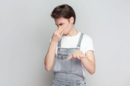 Young brunette man covers nose with hand, smells something awful, pinches nose, frowns in displeasure, sees pile of garbage, wearing denim overalls. Indoor studio shot isolated on gray background.