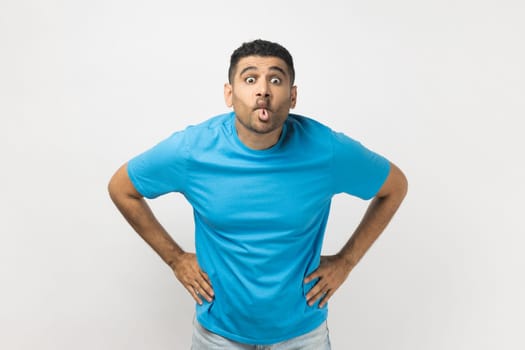 Portrait of funny crazy childish unshaven man wearing blue T- shirt standing with hands on hips, showing tongue out, demonstrates playful behavior. Indoor studio shot isolated on gray background.