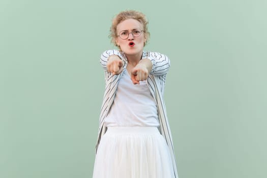 Portrait of angry shocked young adult blonde woman wearing striped shirt and skirt, pointing at you, choosing you, screaming. Indoor studio shot isolated on light green background.