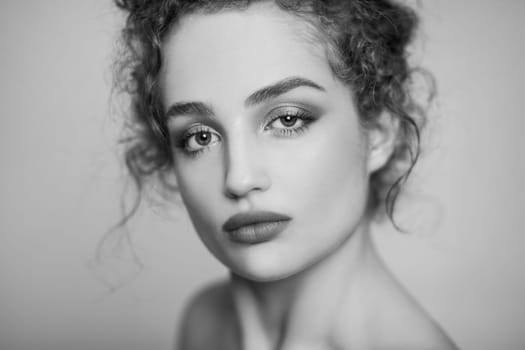 Black and white beauty portrait of young confident woman fashion model with collected dark curly hair and makeup, looking at camera with calm face. Indoor studio shot isolated on gray background.