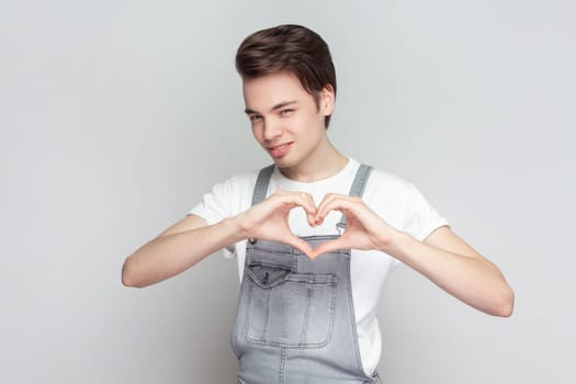 Be my valenetine. Man standing confesses in love, shows heart gesture, smiles broadly, has romantic feelings, seeks lonely hearts, wearing denim overalls. Indoor studio shot isolated on gray background