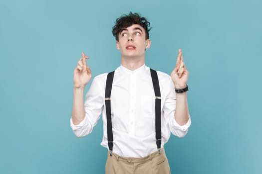 Portrait of hopeful man wearing white shirt and suspender standing with crossed fingers, waiting for miracle, make a wish. Indoor studio shot isolated on blue background.