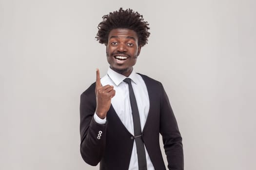 Clever man with Afro hairstyle get new idea, raises fore finger, ready to start work on project, confident in success, wearing white shirt and tuxedo. Indoor studio shot isolated on gray background.