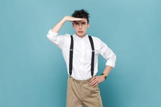 Portrait of serious handsome young man wearing white shirt and suspender standing with palm over forehead, looking far, finding something. Indoor studio shot isolated on blue background.
