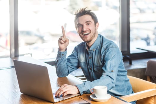 Portrait of excited overjoyed young man freelancer in blue jeans shirt working on laptop and raised finger up, having good idea, looking at camera. Indoor shot near big window, cafe background.