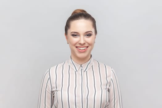 Portrait of attractive joyful cheerful woman wearing striped shirt looking at camera with toothy smile, being in good mood, expressing positiveness. Indoor studio shot isolated on gray background.