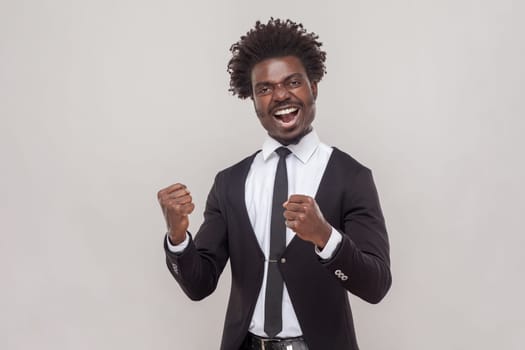 Portrait of overjoyed man with petit goatee clenches fists with joy, feels very happy and lucky, exclaims loudly, wearing white shirt and tuxedo. Indoor studio shot isolated on gray background.