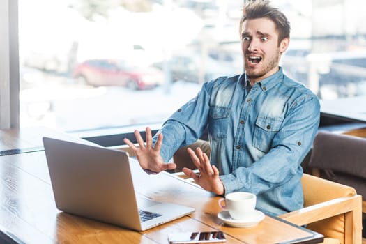 Portrait of shocked panicked scared young man freelancer in blue jeans shirt working on laptop, showing stop gesture to computer screen, looks frighten. Indoor shot near big window, cafe background.