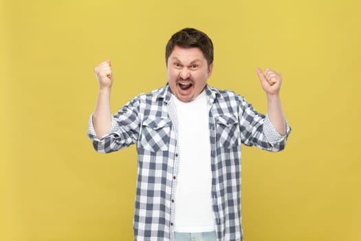 Extremely happy overjoyed middle aged man in casual checkered shirt standing with raised arms and clenched fists, winning lottery, celebrating. Indoor studio shot isolated on yellow background.
