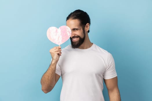 Portrait of man with beard wearing white T-shirt holding and covering eye with paper pink heart on stick and smiling to camera, sharing love. Indoor studio shot isolated on blue background.