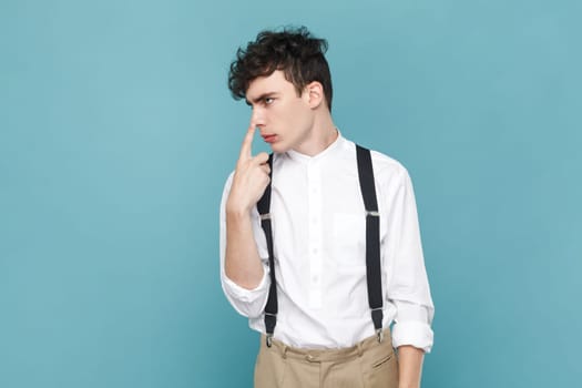 Portrait of man wearing white shirt and suspender, standing, touching his nose and showing lie gesture, suspecting falsehood, dishonest talk. Indoor studio shot isolated on blue background.