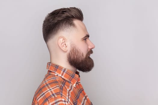 Side view portrait of serious strict bossy bearded man standing looking ahead, expressing confidence, wearing orange checkered shirt. Indoor studio shot isolated on gray background.