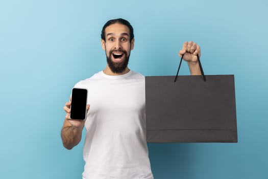 Portrait of astonished man with beard wearing white T-shirt shocked with purchases holding paper shopping bags and smart phone with empty display. Indoor studio shot isolated on blue background.