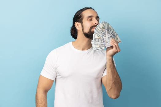 Portrait of rich man with beard wearing white T-shirt holding big fan of money, smelling dollars banknotes with pleasure. Indoor studio shot isolated on blue background.