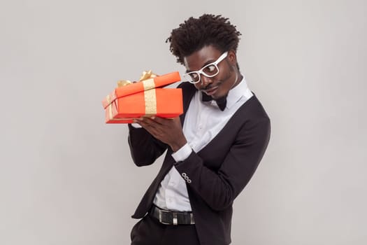 Portrait of curious handsome young adult man in glasses standing with red gift box, looking inside, wearing white shirt and tuxedo. Indoor studio shot isolated on gray background.