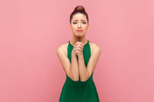 Portrait of sad desperate woman with bun hairstyle standing keeps hands in praying gesture, pleading, apologizing, wearing green dress. Indoor studio shot isolated on pink background.