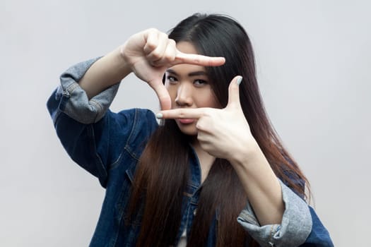 Portrait of serious concentrated attractive brunette woman in blue denim jacket standing looking at camera and gesturing finger frame. Indoor studio shot isolated on gray background.