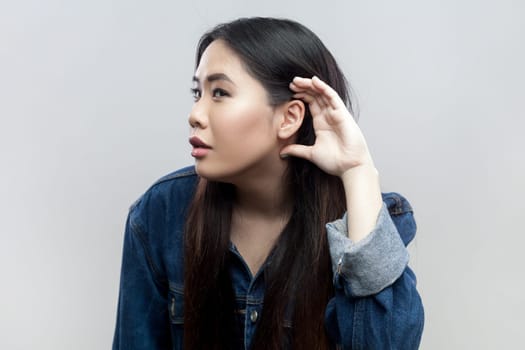 Portrait of brunette woman in blue denim jacket standing holding hand near ear, listening to interesting talks and private secrets, spying. Indoor studio shot isolated on gray background.
