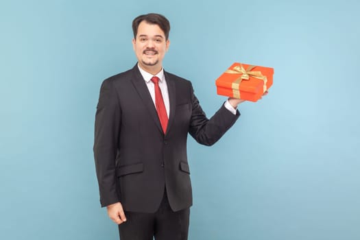 Portrait of satisfied cheerful man with mustache standing holding red gift box, preparing present to his wife, wearing black suit with red tie. Indoor studio shot isolated on light blue background.