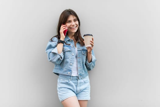Portrait of happy satisfied attractive woman wearing denim jacket having rest at work, drinking coffee to go and talking with boyfriend on cell phone. Indoor studio shot isolated on gray background.
