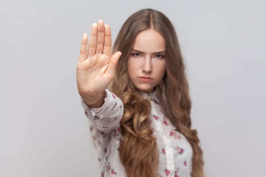 Portrait of confident serious attractive young adult woman with wavy blond hair showing block stop gesture, set back bullying and violence. Indoor studio shot isolated on gray background.