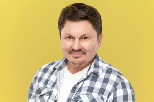 Portrait of smiling middle aged man in casual checkered shirt with beard and mustache looking at camera with positive expression. Indoor studio shot isolated on yellow background.