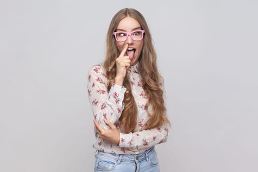 Portrait of funny woman in glasses with wavy blond hair putting finger into his nose, fooling around, bad habits, disrespectful behavior. Indoor studio shot isolated on gray background.