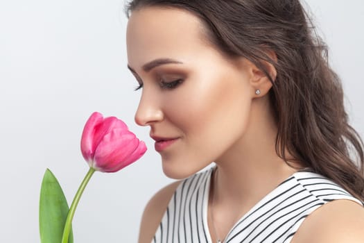 Closeup portrait of romantic dreaming brunette woman standing holding pink tulip, enjoying aromat of flower, wearing striped dress. Indoor studio shot isolated on gray background.
