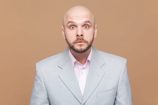 Portrait of shocked startled bald bearded man stares bugged eyes at camera hears big news, feels very surprised, wearing gray jacket. Indoor studio shot isolated on brown background.