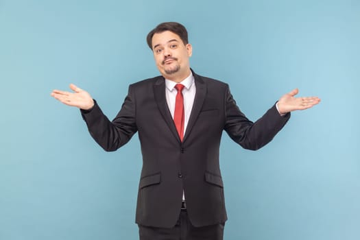 Portrait of confused uncertain man with mustache with spread hands, looking at camera, shrugging shoulders, wearing black suit with red tie. Indoor studio shot isolated on light blue background.