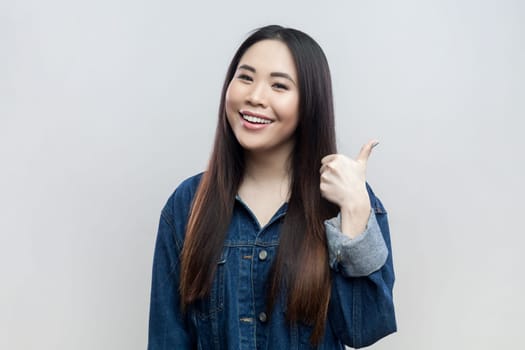 Portrait of happy smiling attractive brunette woman in blue denim jacket standing showing thumb up, like gesture, recommend service. Indoor studio shot isolated on gray background.