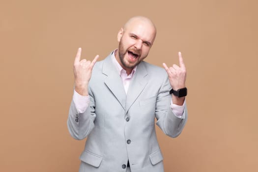 Portrait of bald bearded man rocker enjoys positive vibes, listens rock n roll, cool music in club, shows horn gesture, wearing gray jacket. Indoor studio shot isolated on brown background.